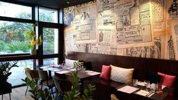 A restaurant with a wall of vintage newspaper clippings, large windows, and indoor plants.
