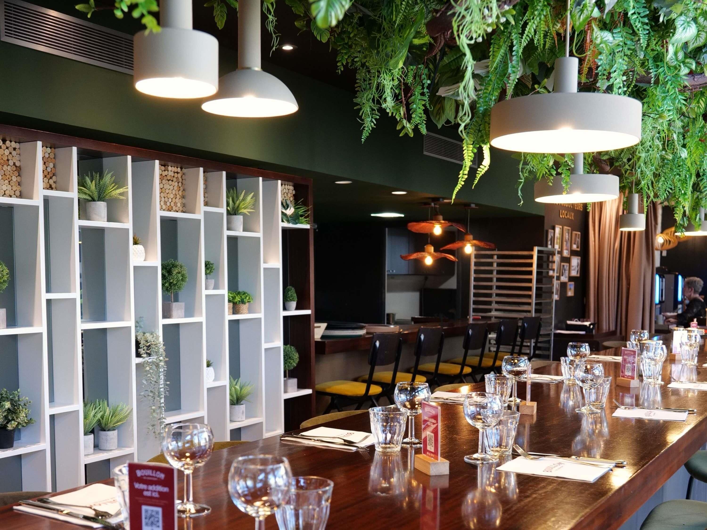 Un restaurante con una larga mesa de madera dispuesta para comer, rodeado de estanterías blancas con plantas y luces colgantes.