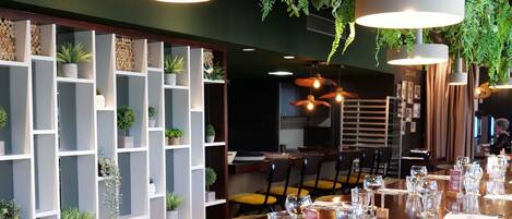 Un restaurante con una larga mesa de madera dispuesta para comer, rodeado de estanterías blancas con plantas y luces colgantes.