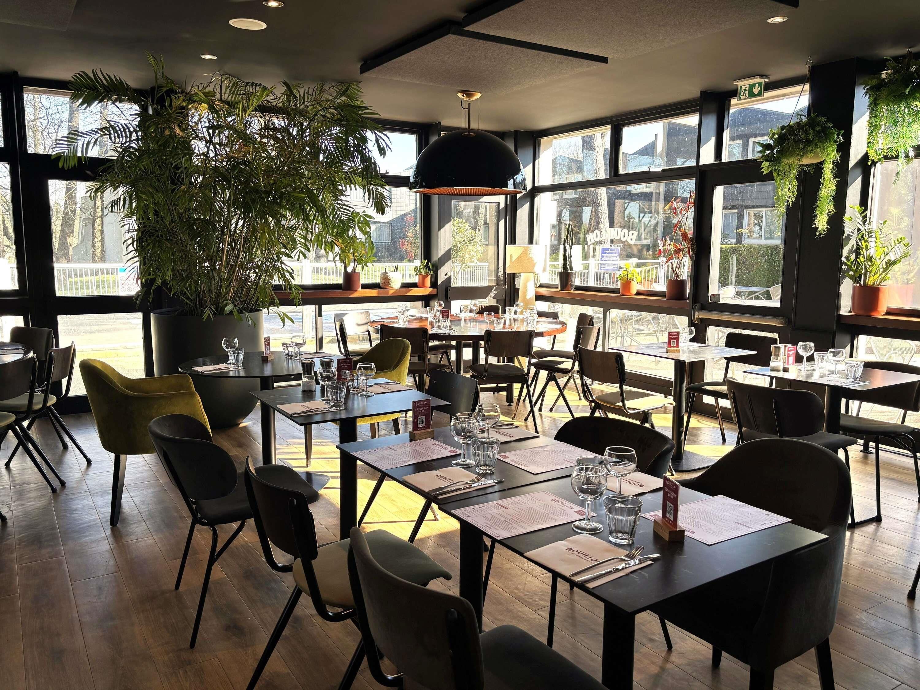 A restaurant with tables set for dining, large windows, and indoor plants.