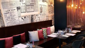 A restaurant with a wall of vintage newspapers, a long dining table with chairs, and a warm light fixture.