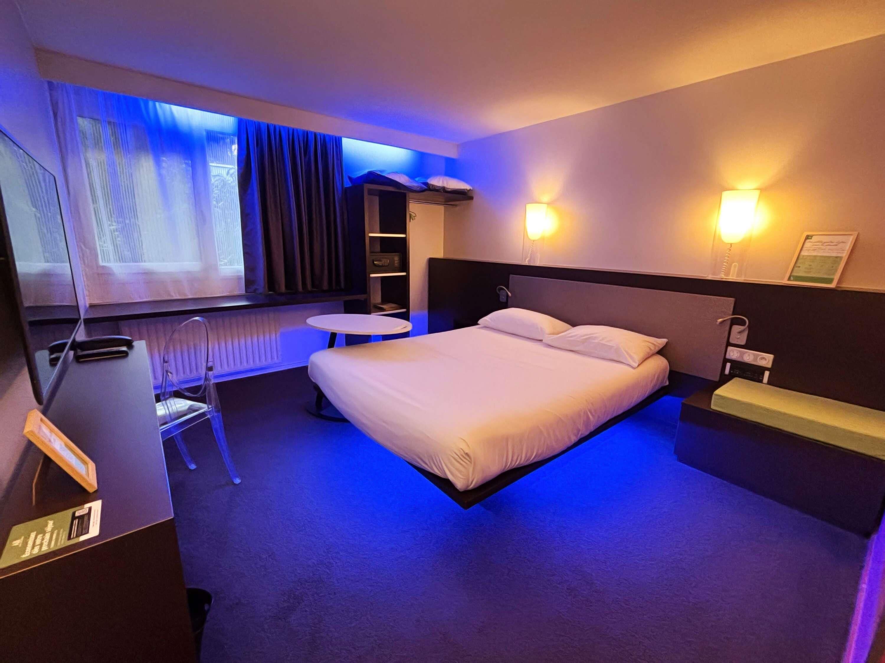 A hotel room with a bed, bedside table, lamp, and a small desk with a chair.