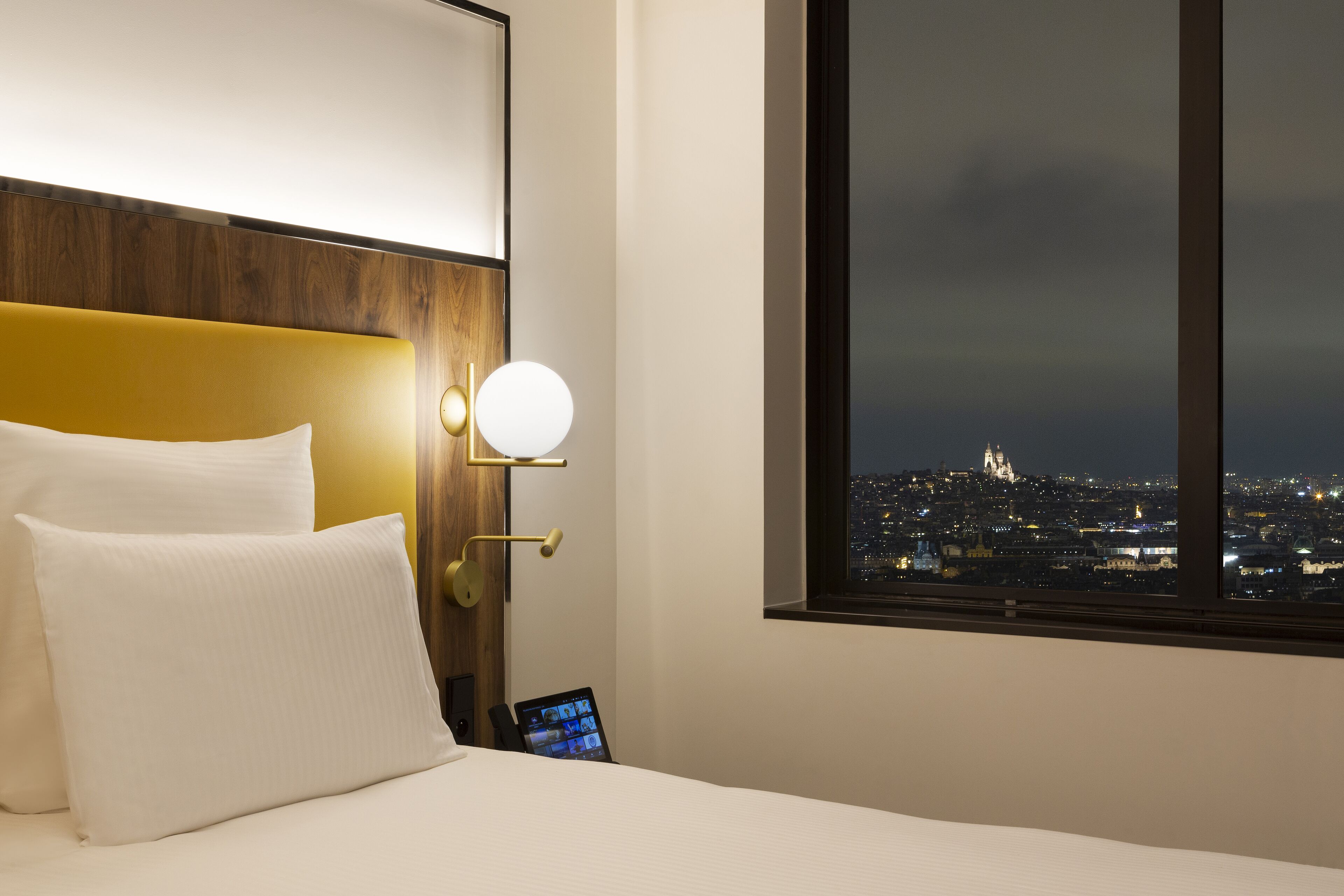 A hotel room with a bed, a nightstand, a tablet, a lamp, and a window with a city view.