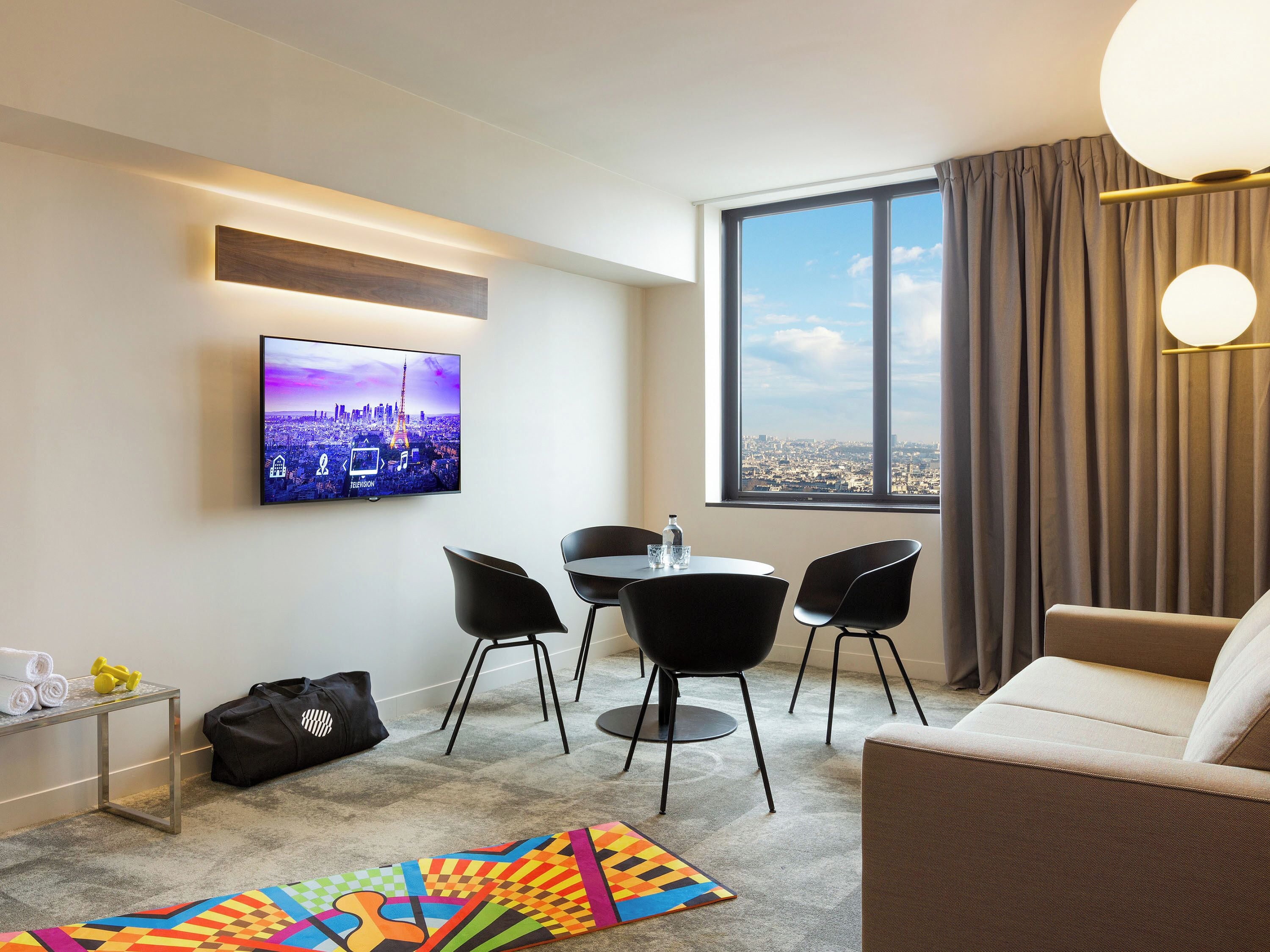 A modern hotel room with a cityscape view, a flat-screen TV, a dining area with chairs, a sofa, and a colorful rug.
