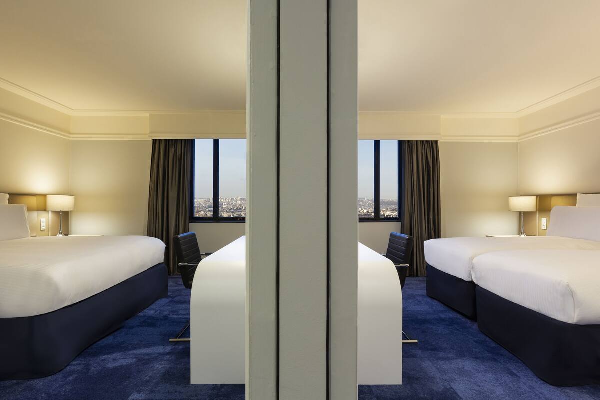 A hotel room with two beds, a desk, and a view of the city.