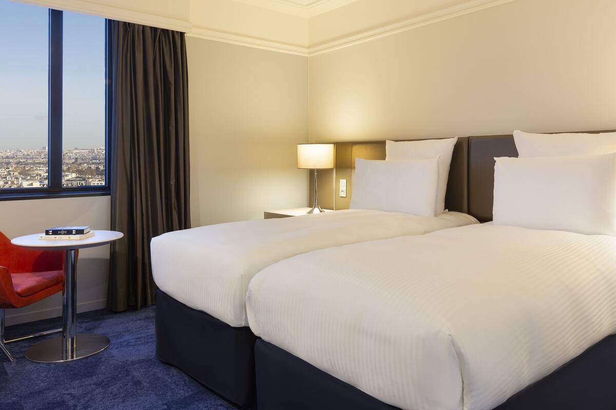 A hotel room with two beds, a small table, a chair, a lamp, and a view of the city.