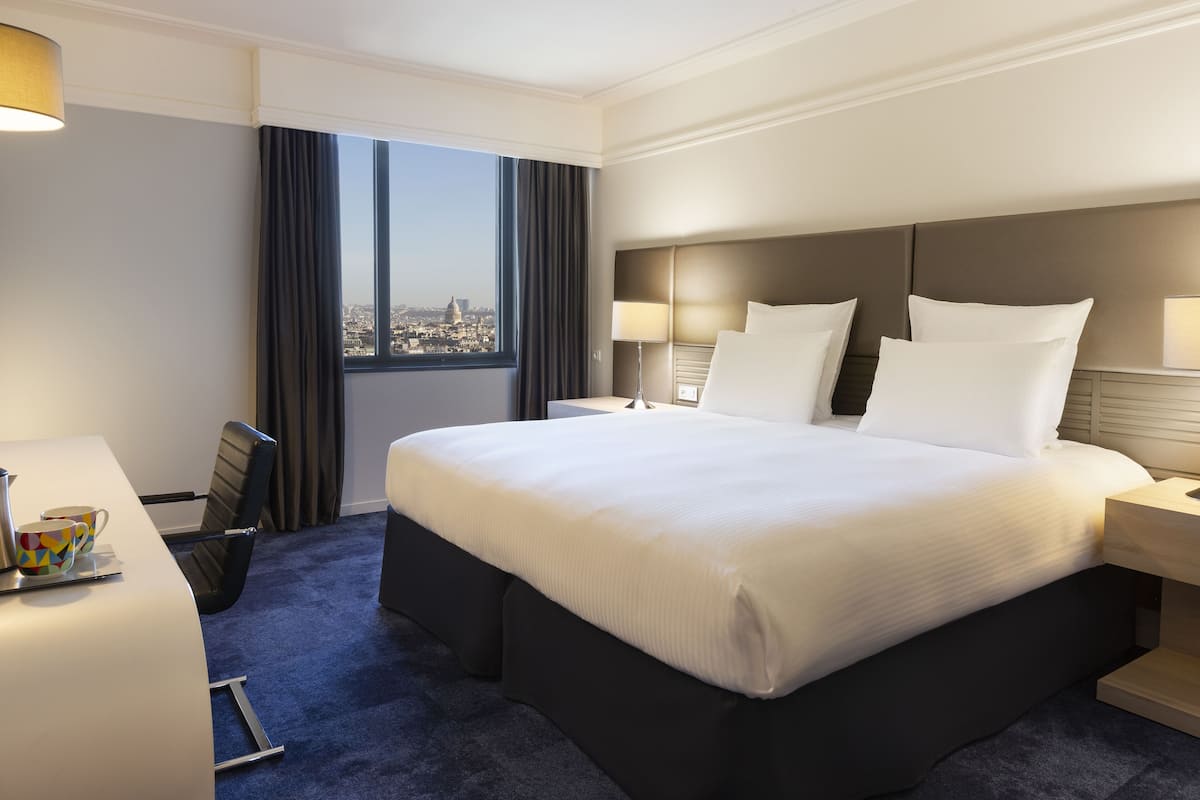 A hotel room with a large bed, a desk, a chair, a lamp, and a view of the city through the window.
