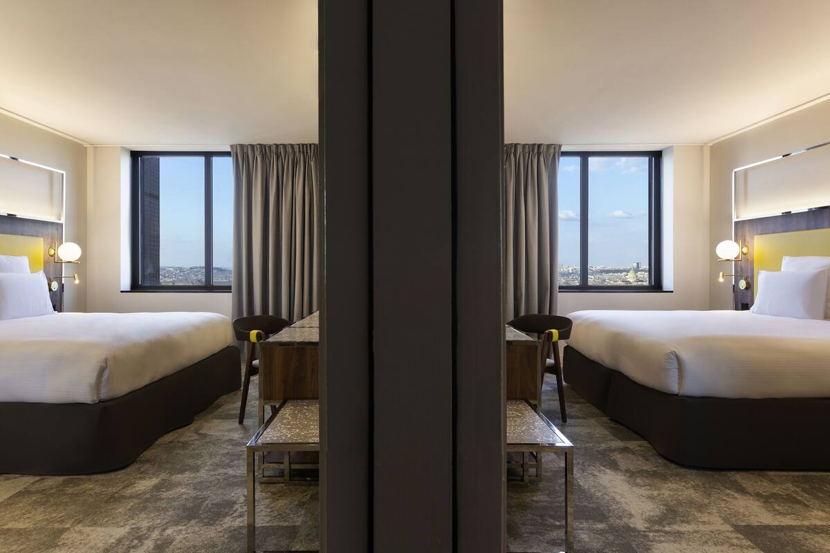 A hotel room with two beds, a dining table, and large windows offering a city view.