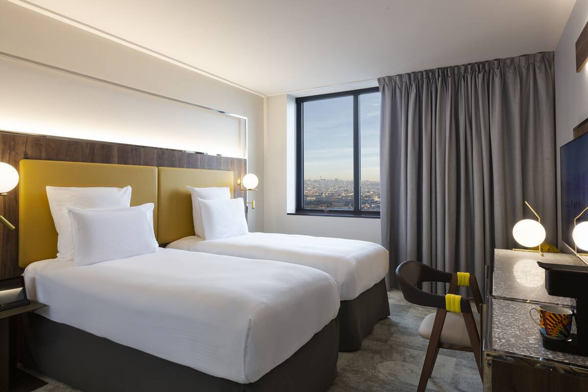 A hotel room with two beds, a desk with a chair, a TV, and a large window with a city view.