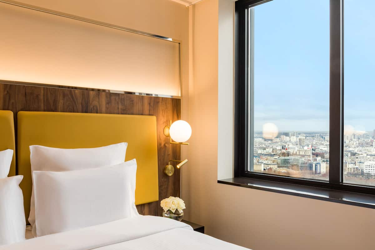A hotel room with a large bed, two pillows, a nightstand with a vase of flowers, a wall-mounted light, and a window with a city view.