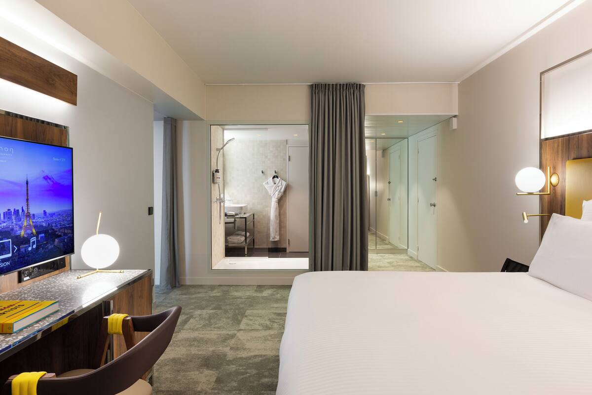 A hotel room with a bed, a desk with a flat-screen TV, a chair, and a view of the bathroom.