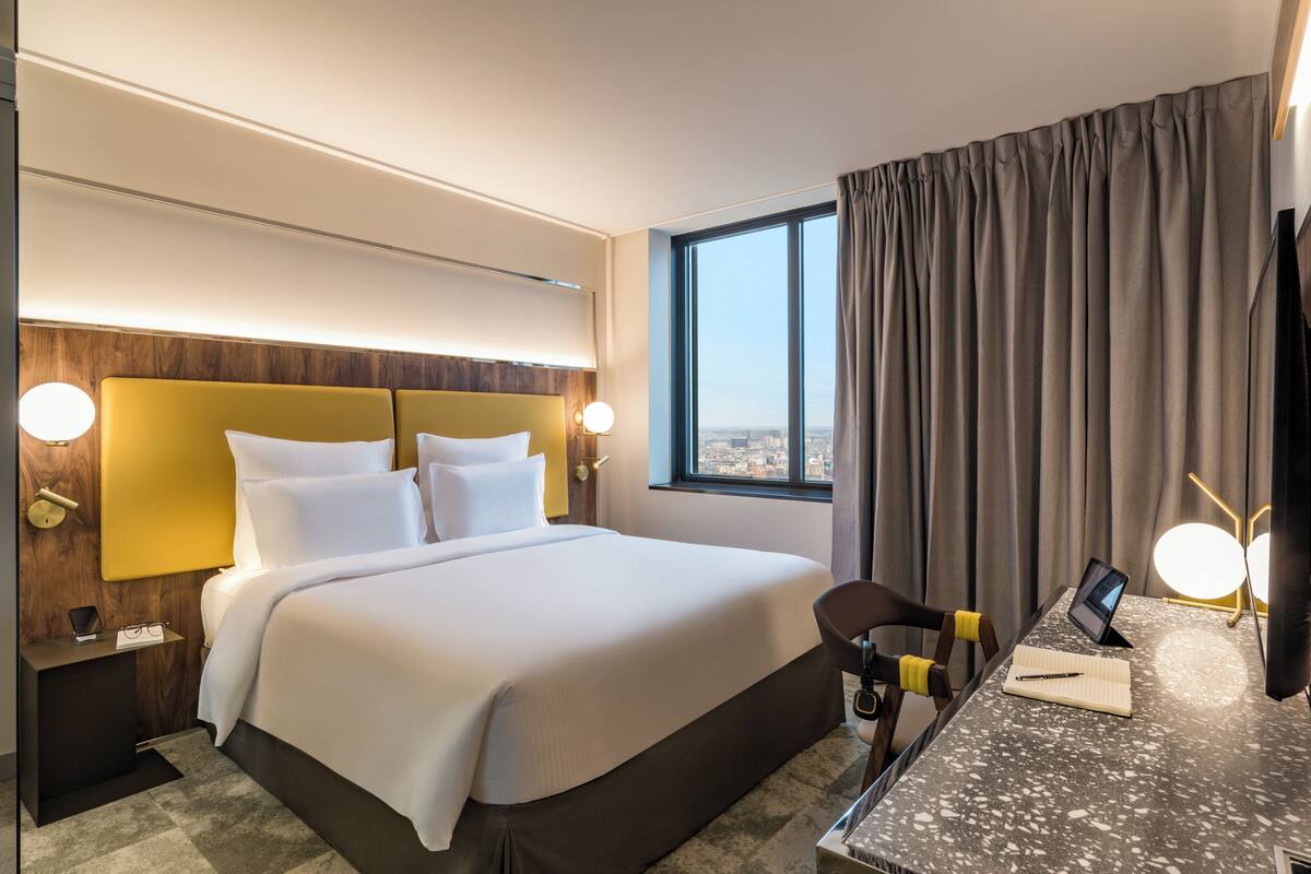 A hotel room with a large bed, a desk with a chair, a window with curtains, and a view of the city.