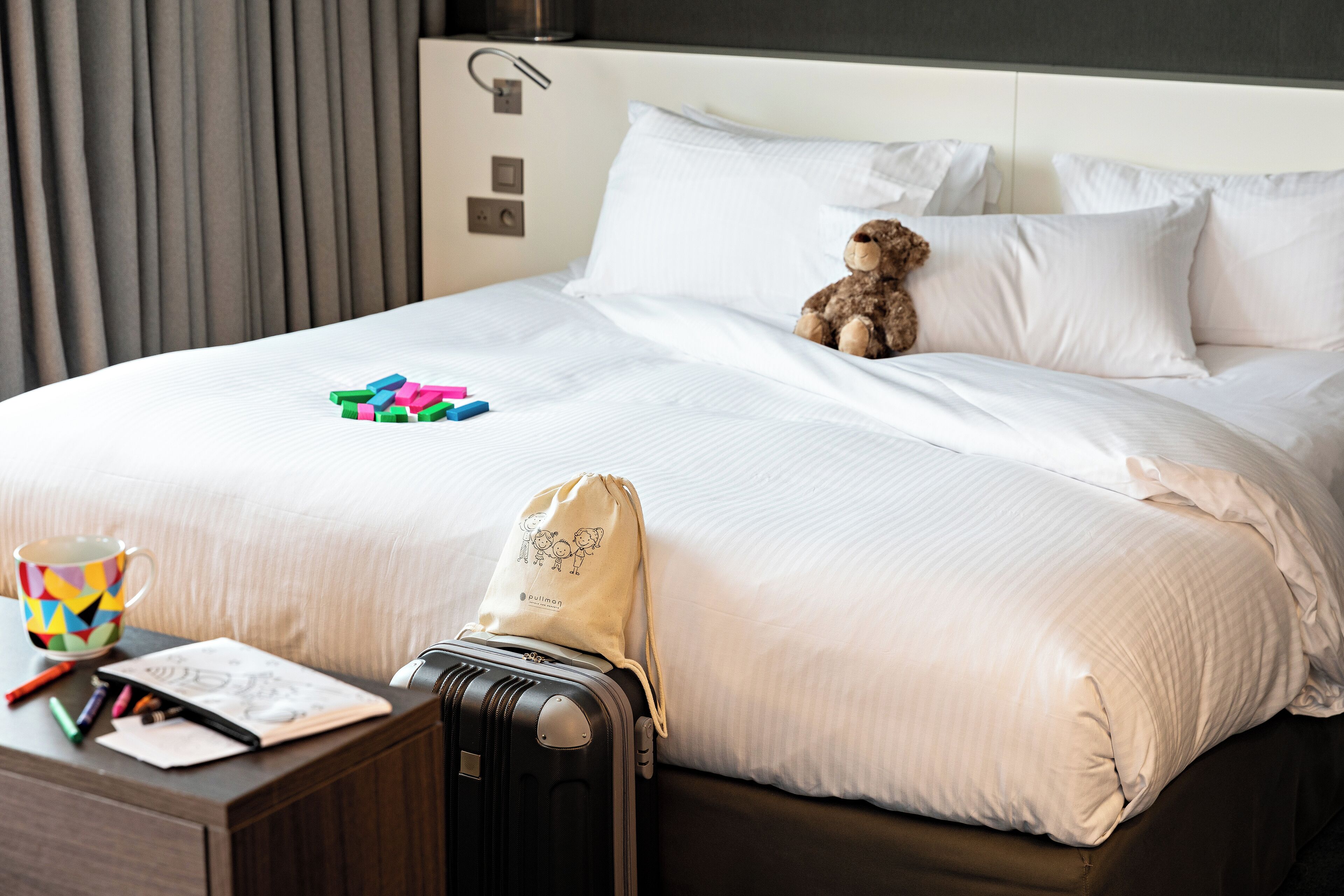 A neatly made bed with a teddy bear, a colorful mug, a notebook, and a suitcase on a wooden nightstand.