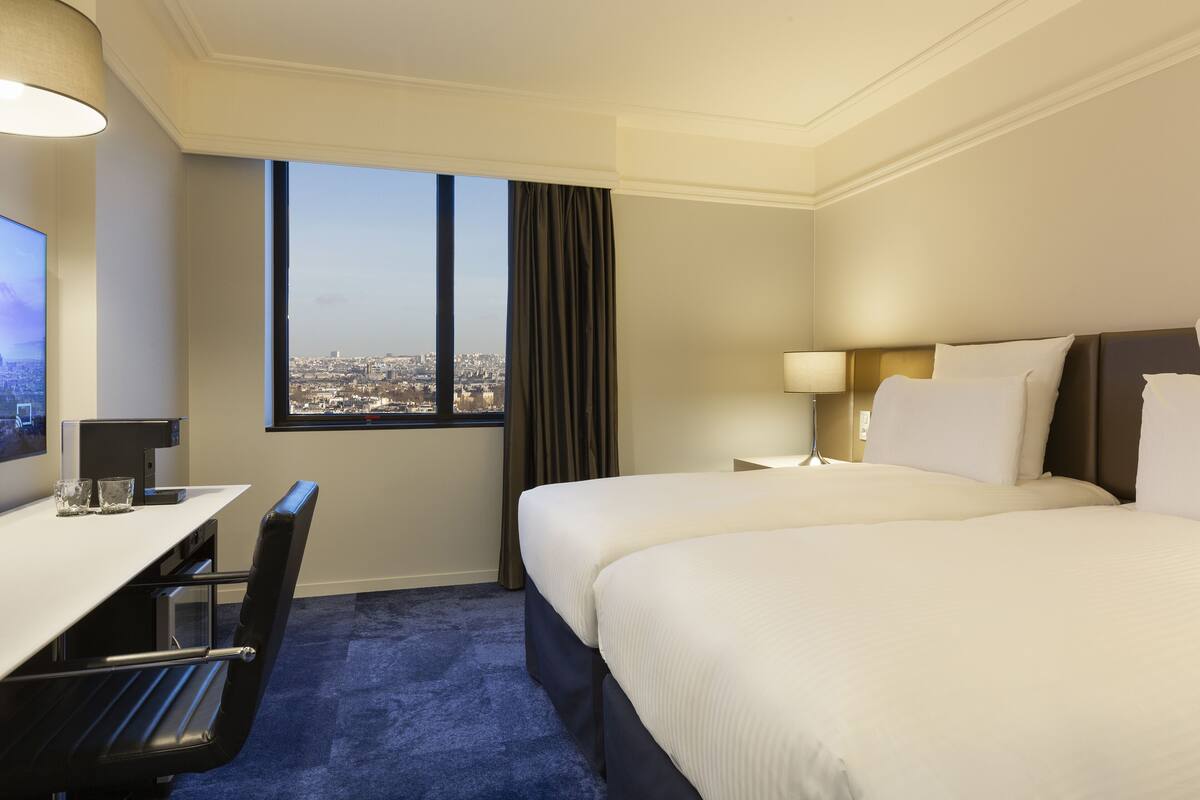 A hotel room with a large bed, a desk with a chair, a TV, and a window with a city view.