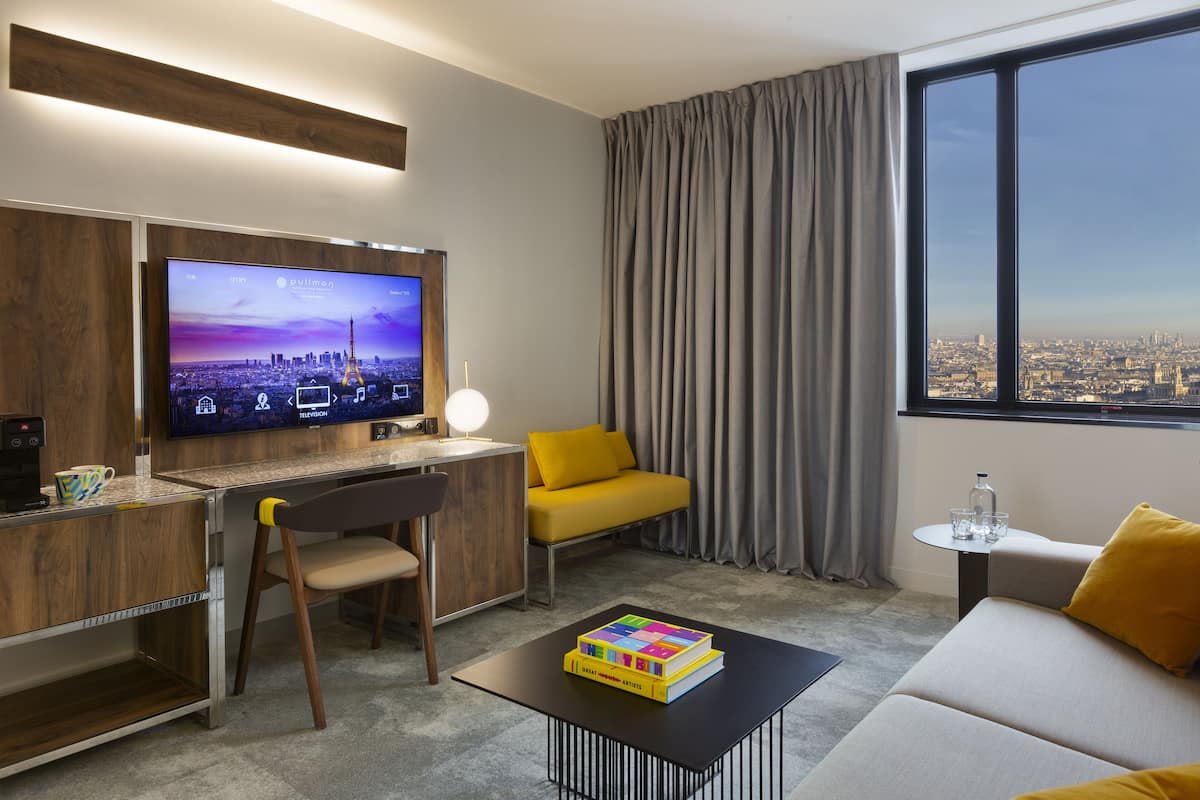A modern hotel room with a large TV, a desk, a sofa, and a view of the city.