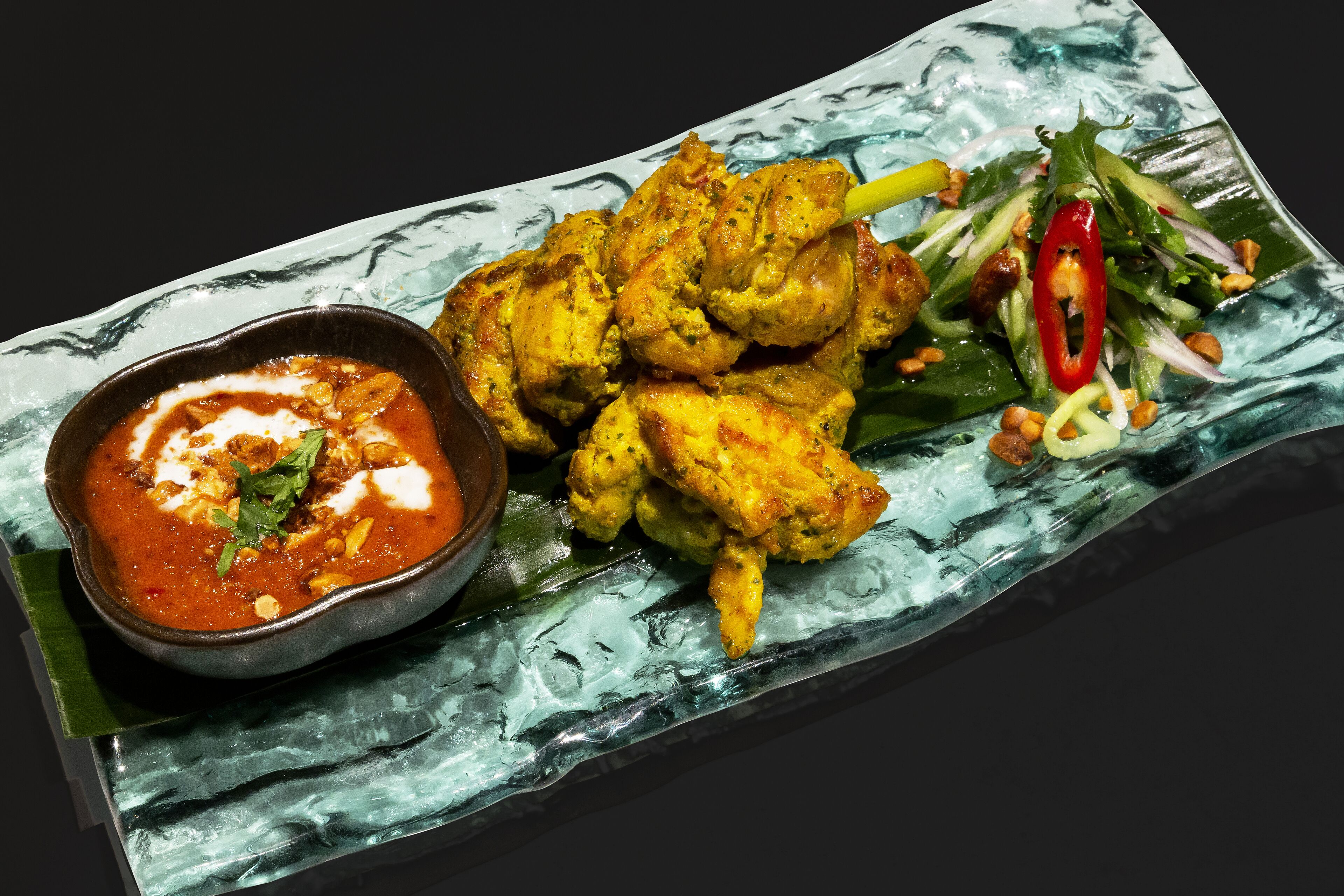 En rektangulär tallrik med grillade kycklingspett, en skål med röd sås och en sallad som tillbehör, bestående av grönsaker och en röd chili.