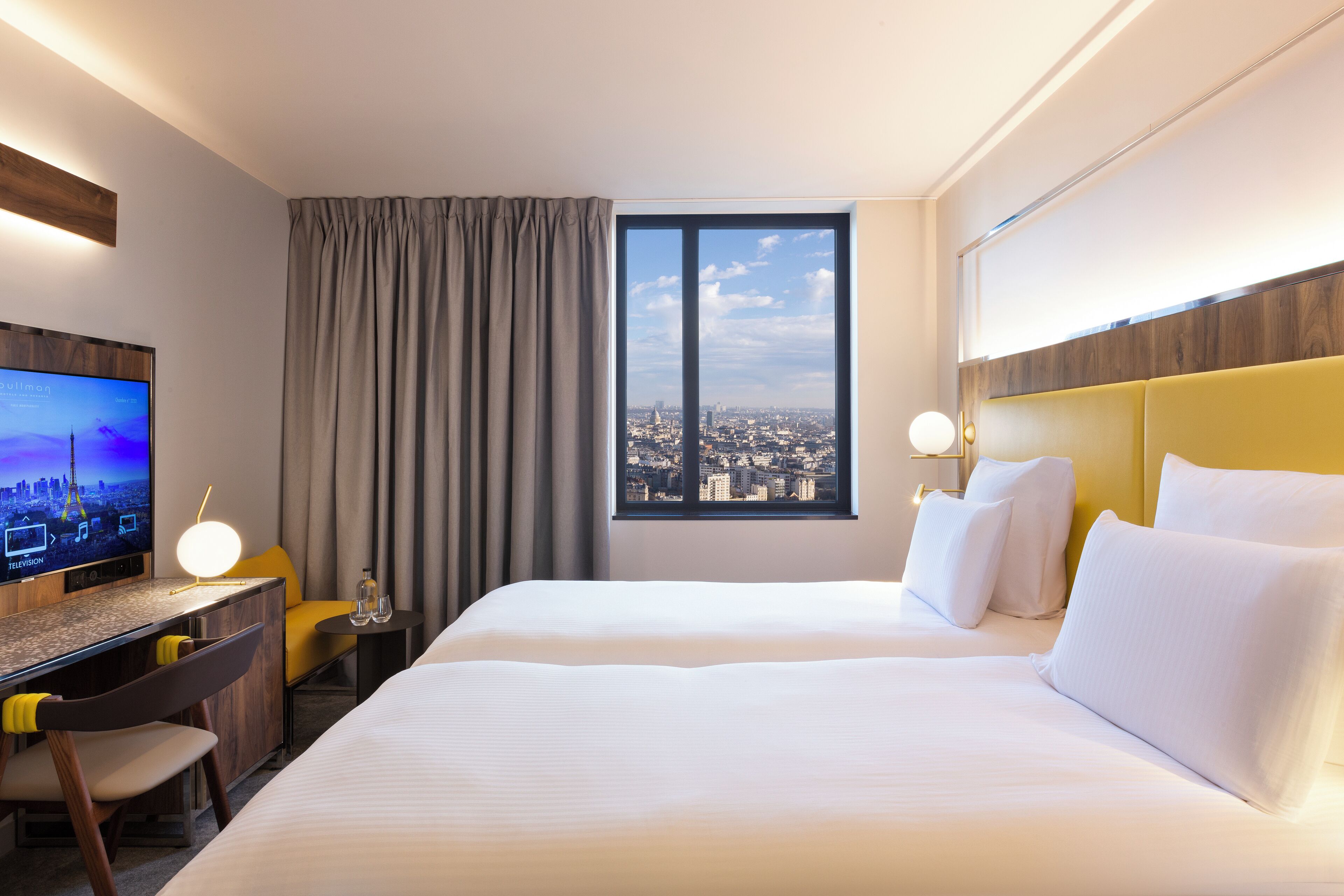 A hotel room with a bed, a desk with a TV, a chair, a window with a city view, and a headboard.