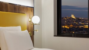 A hotel room with a bed, a round wall-mounted light, and a window with a cityscape view. - Pullman Paris Montparnasse Hotel (Paris)