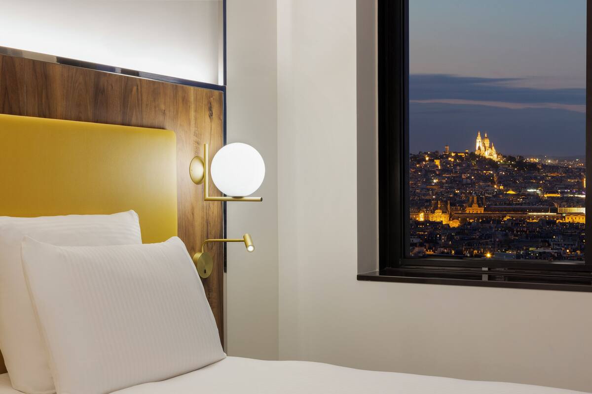 A hotel room with a bed, a round wall-mounted light, and a window with a cityscape view.