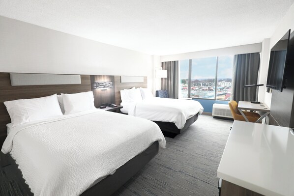 A hotel room with two beds, a desk, a chair, a TV, and a large window with a city view.