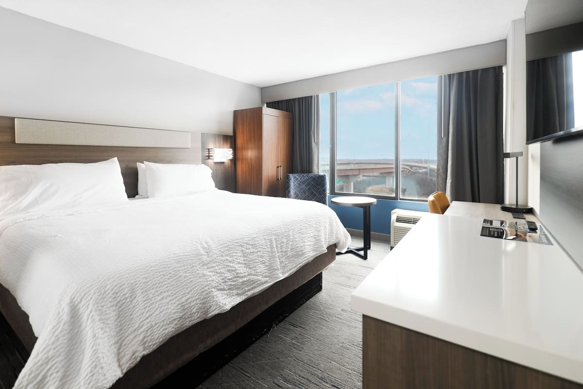 A hotel room with a large bed, a desk, a chair, a TV, and a view of the city.