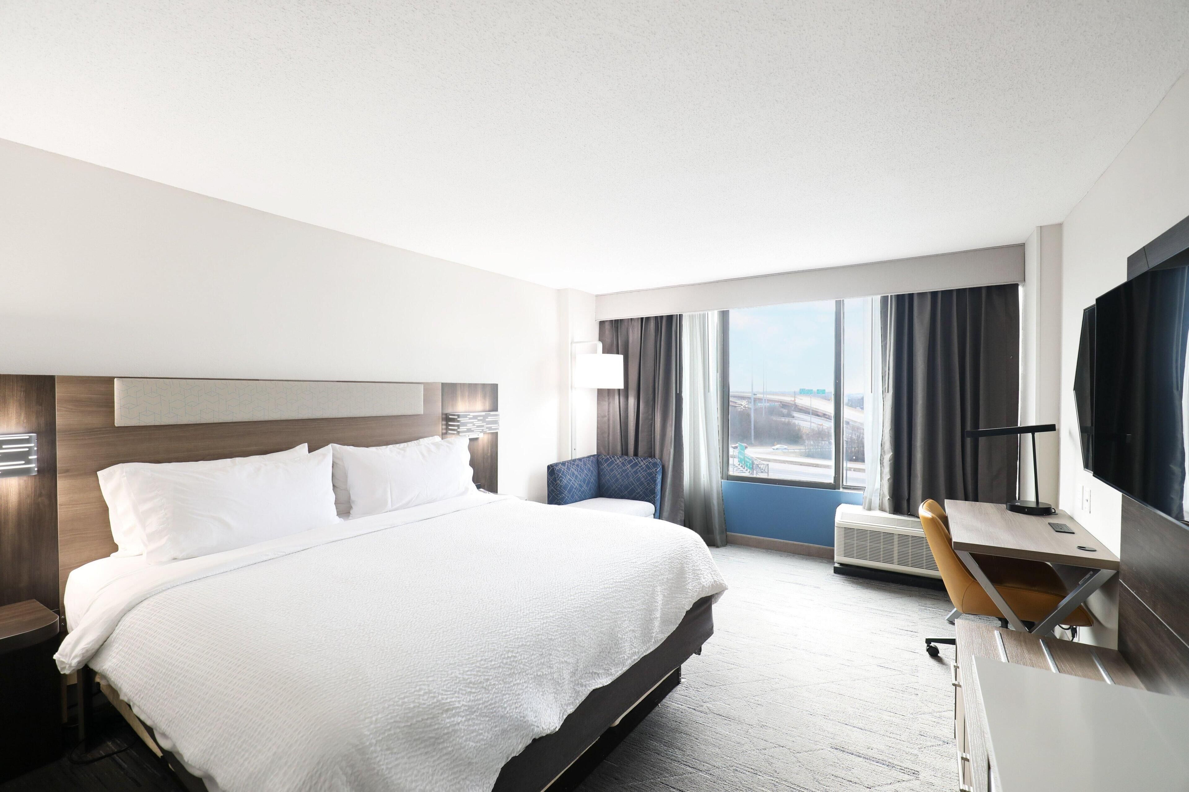 Quarto de hotel com uma cama grande, uma escrivaninha com cadeira, televisão e janela com cortinas.