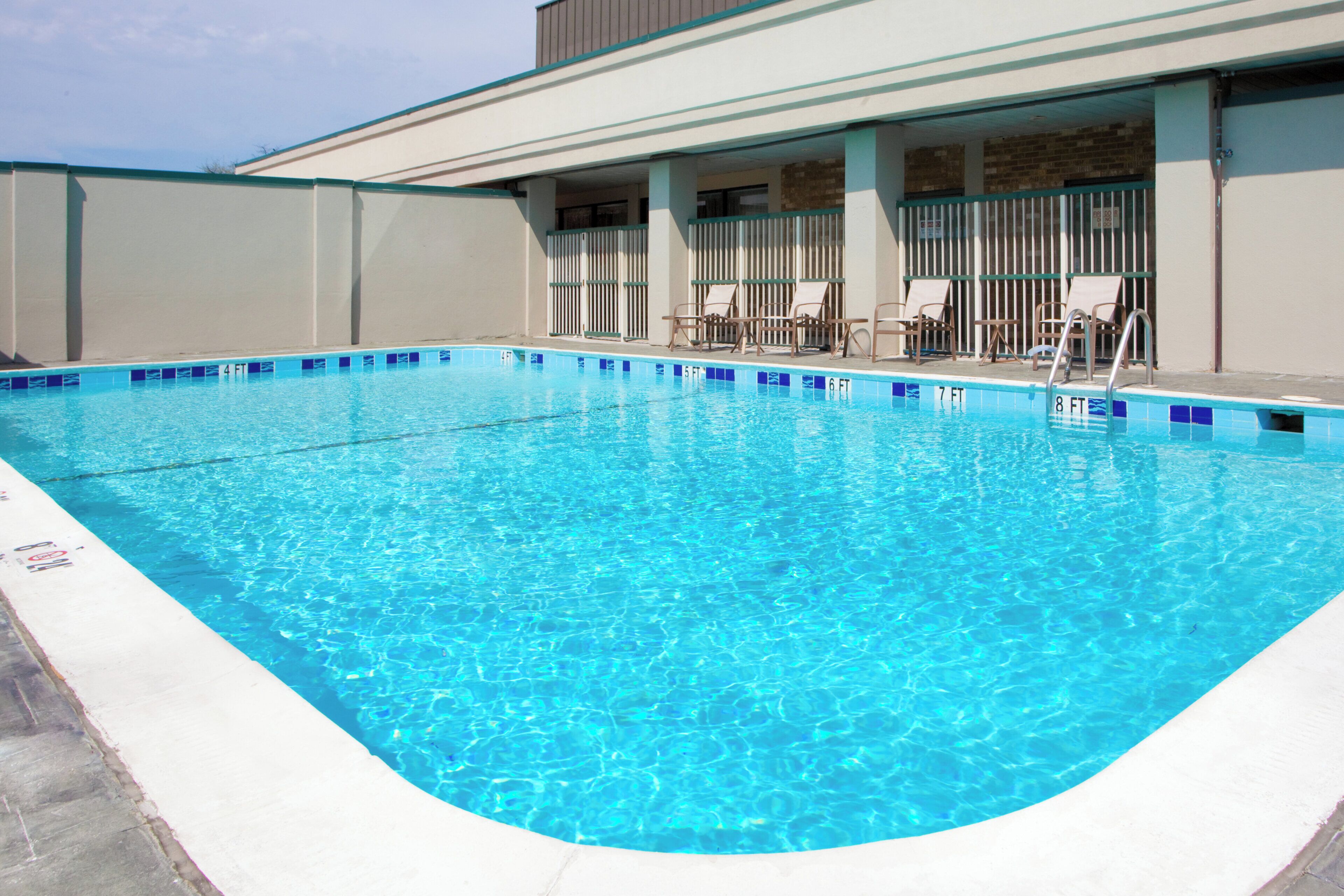 Una piscina rectangular con agua azul cristalina, rodeada por un muro bajo y una zona de estar cubierta con sillas.