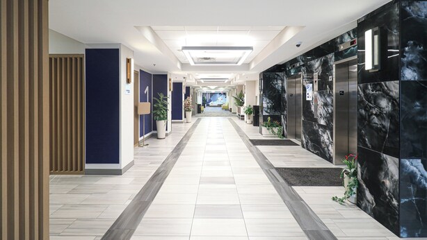 A modern hotel corridor with a central walkway, potted plants, and elevators on either side. - Holiday Inn Express Washington DC SW - Springfield by IHG (Springfield)