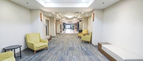 A hotel corridor with a carpeted floor, two yellow armchairs, a small table, and a bench.