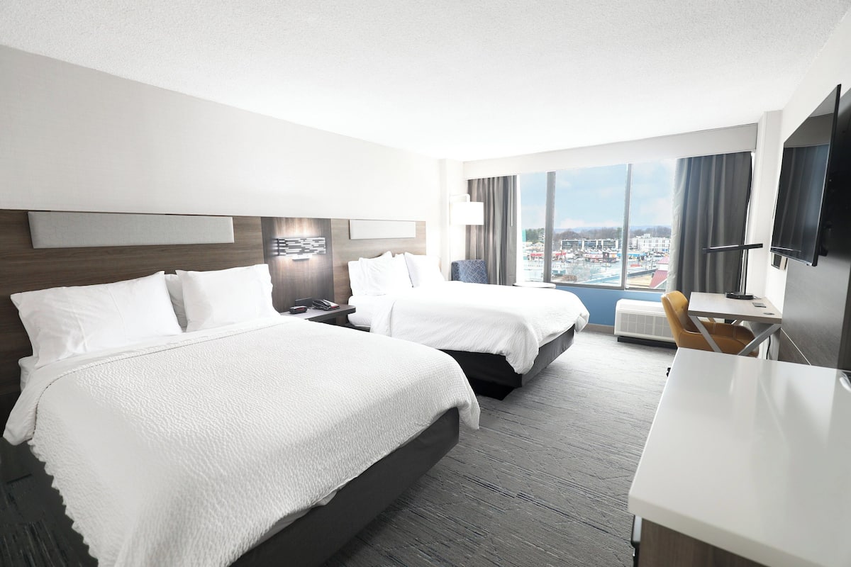 A hotel room with two beds, a desk, a chair, a TV, and a large window with a city view.