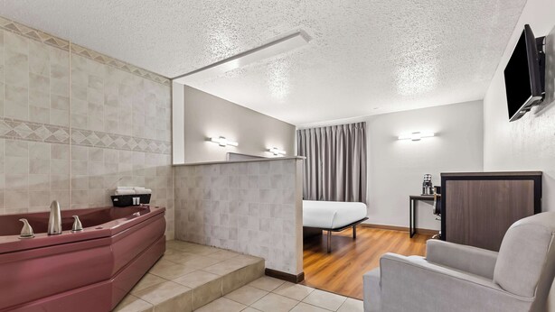 A modern bathroom with a red bathtub, a white sink, and a shower. There is a bed with a white mattress and a grey curtain in the adjacent room. - Red Lion Inn & Suites Pontoon Beach (Granite City)