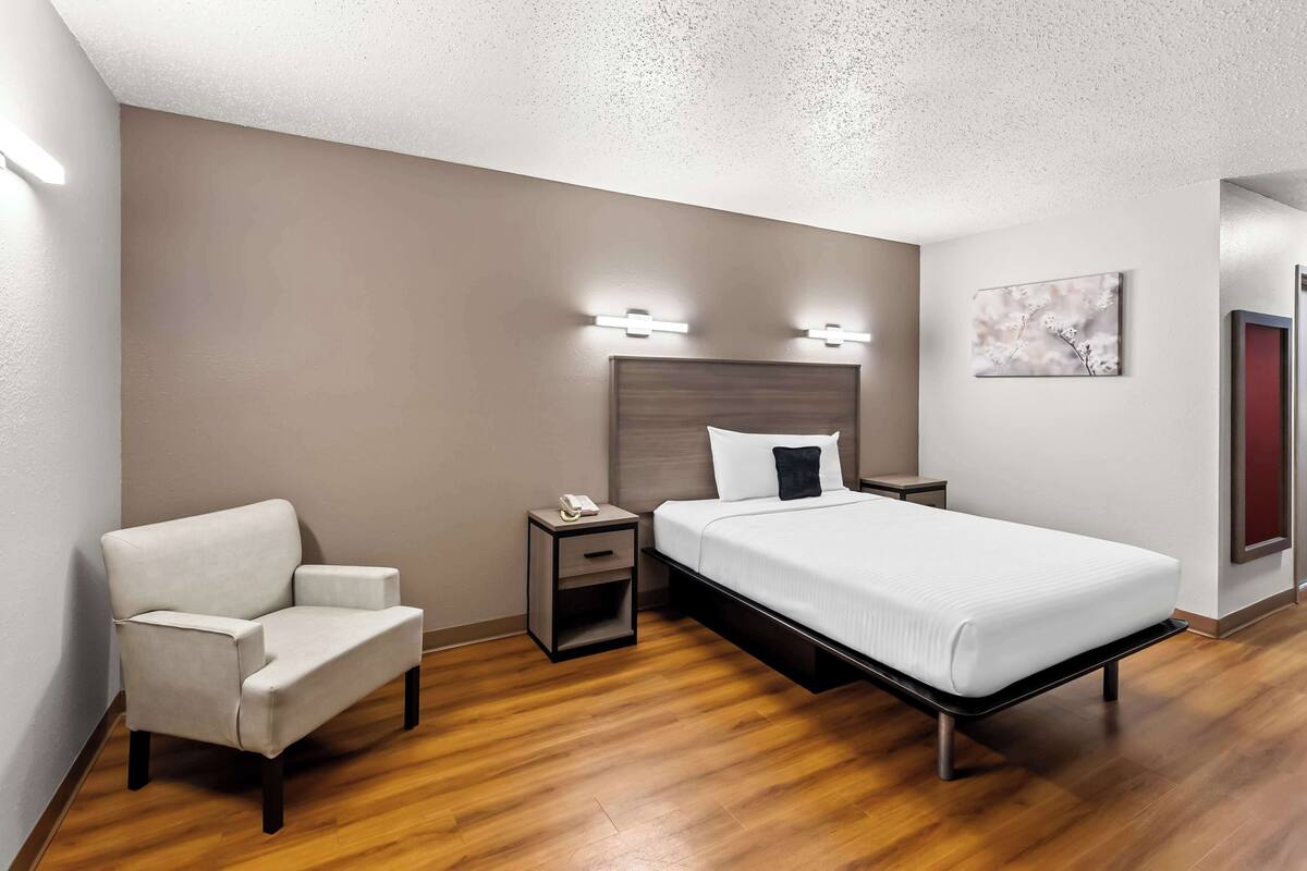 Une chambre d’hôtel avec un lit, une chaise, une table de chevet et un luminaire fixé au mur.