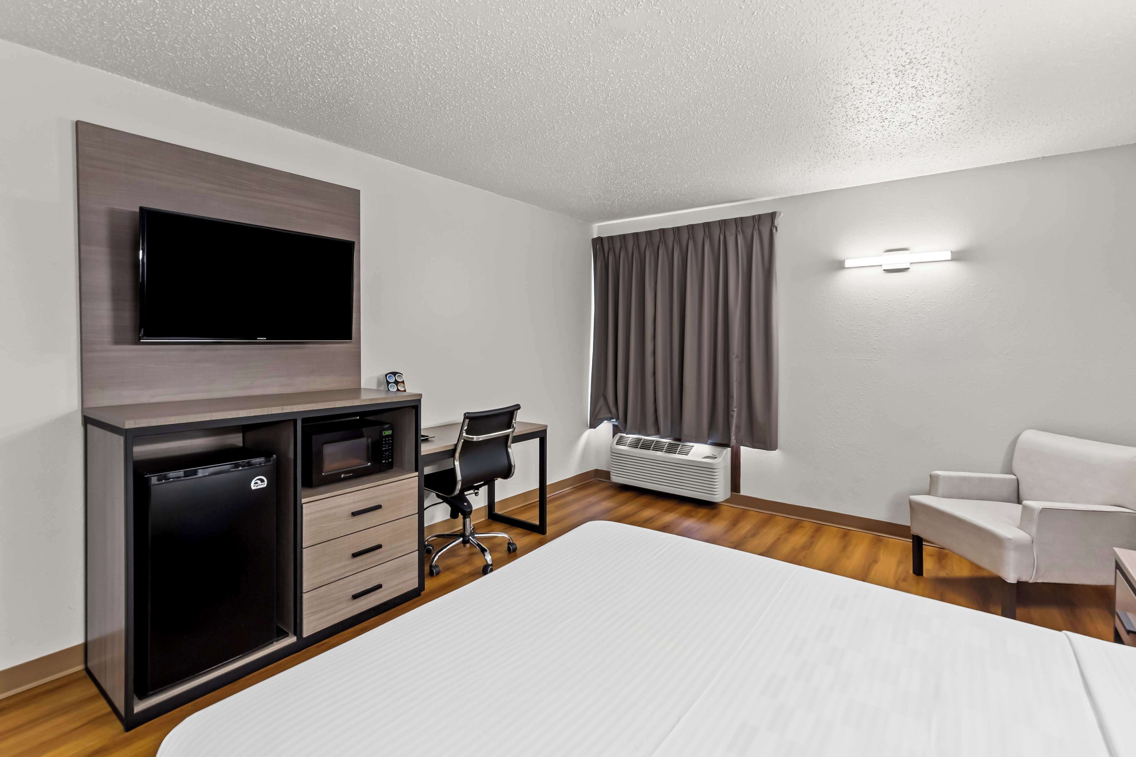 A hotel room with a bed, a desk with a chair, a TV mounted on the wall, a mini-fridge, and an air conditioning unit.