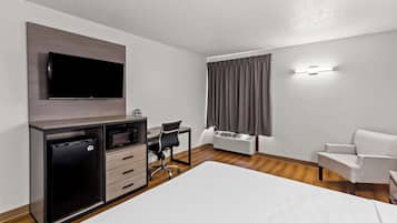A hotel room with a bed, a desk with a chair, a TV mounted on the wall, a mini-fridge, and an air conditioning unit.
