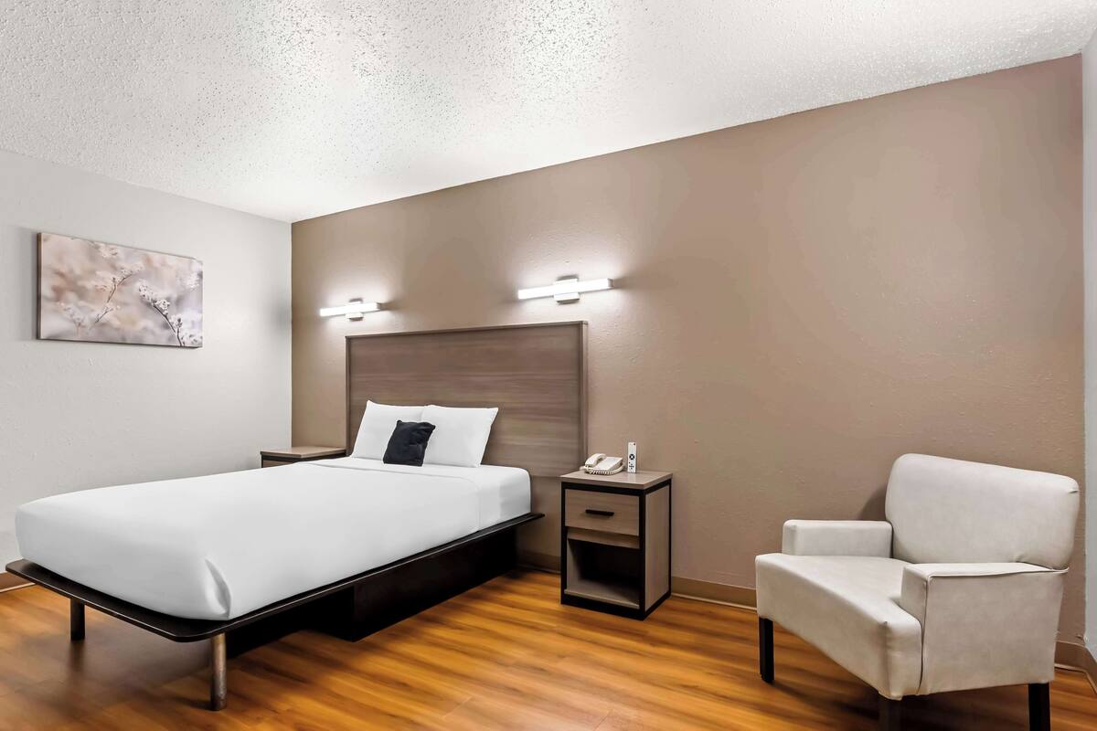 Une chambre d’hôtel moderne équipée d’un lit, d’une table de chevet, d’une chaise et d’un luminaire fixé au mur.