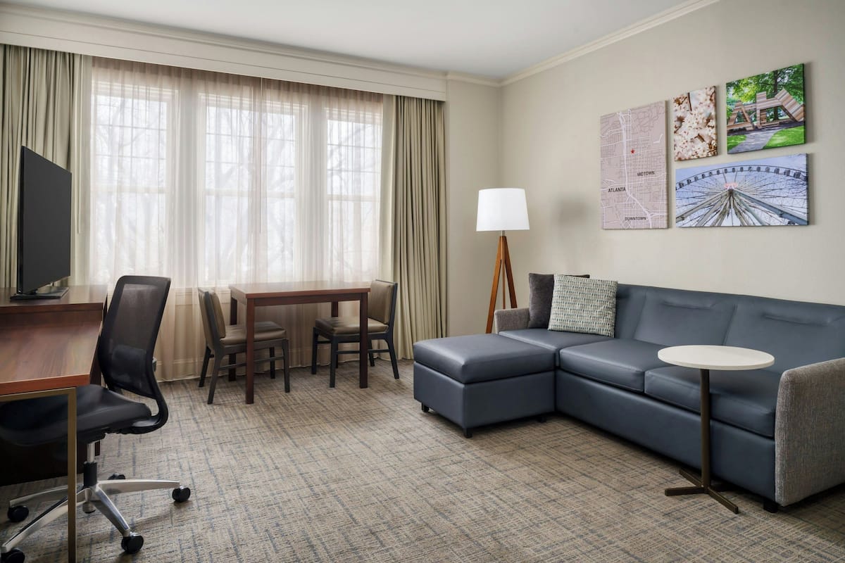 Une chambre d’hôtel comprenant un canapé d’angle, une petite table ronde, un bureau avec une chaise, une télévision et une lampe. Trois œuvres d’art murales représentent des cartes et une grande roue.
