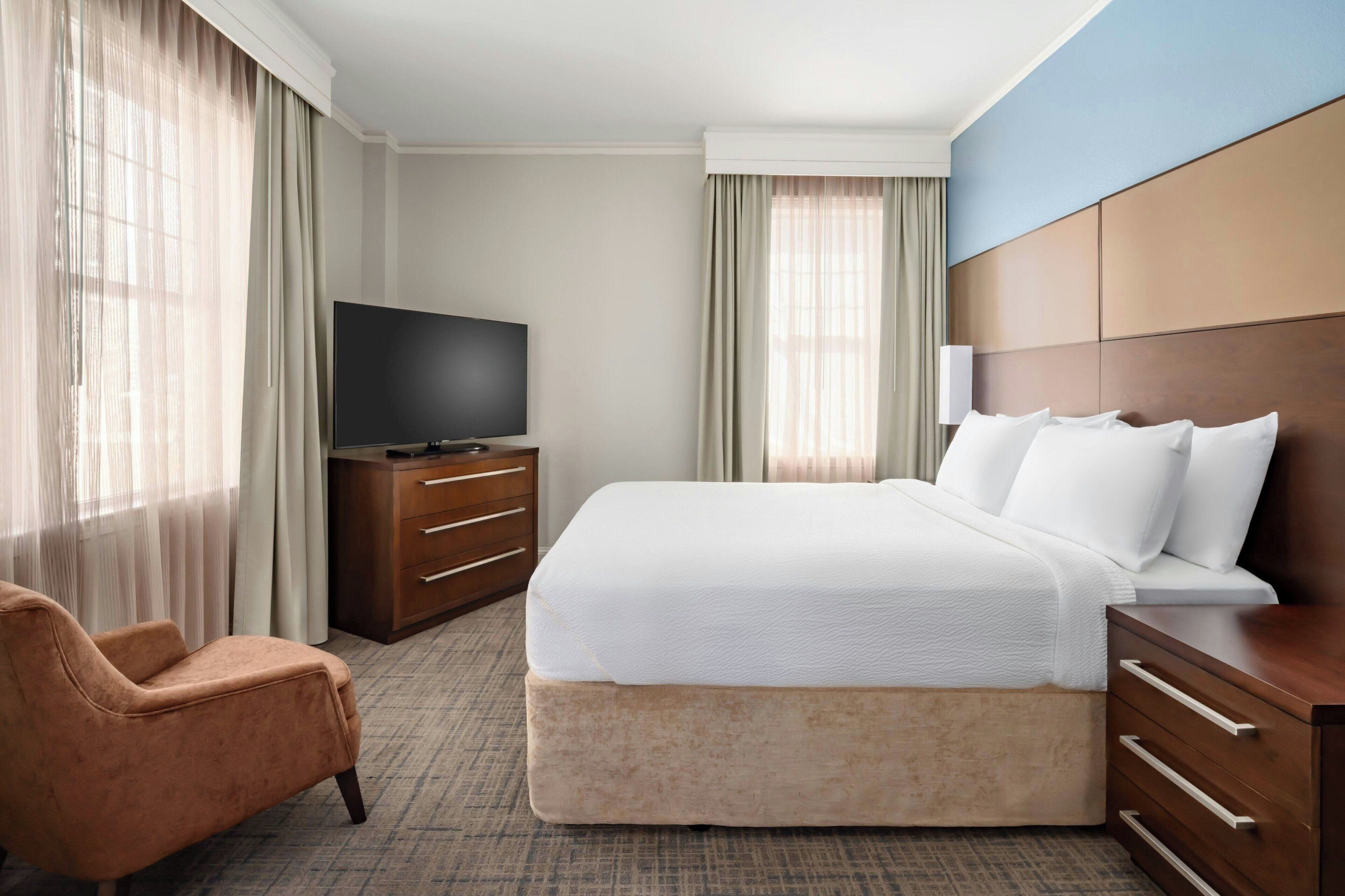 Una habitación de hotel con cama, televisión, silla, cómoda y cortinas.