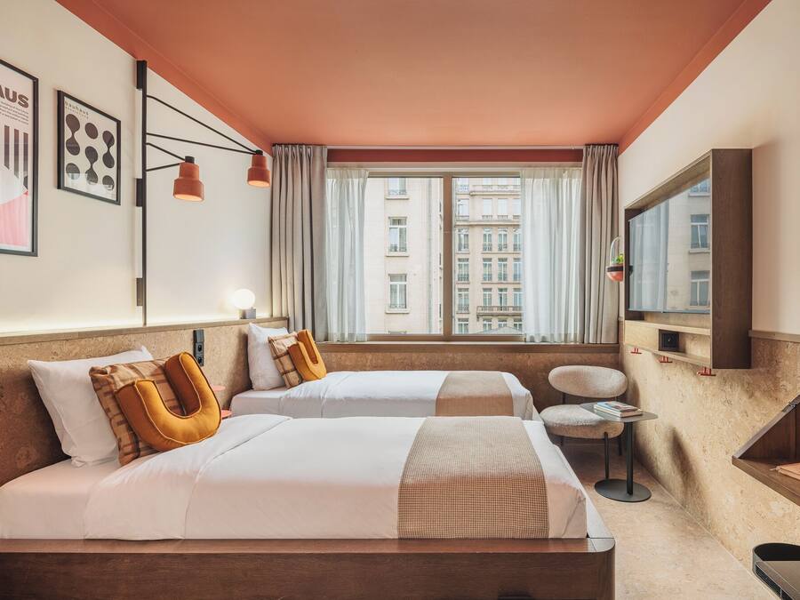 Habitación de hotel con dos camas, una mesita, una silla, televisor y ventana con vista a edificios.