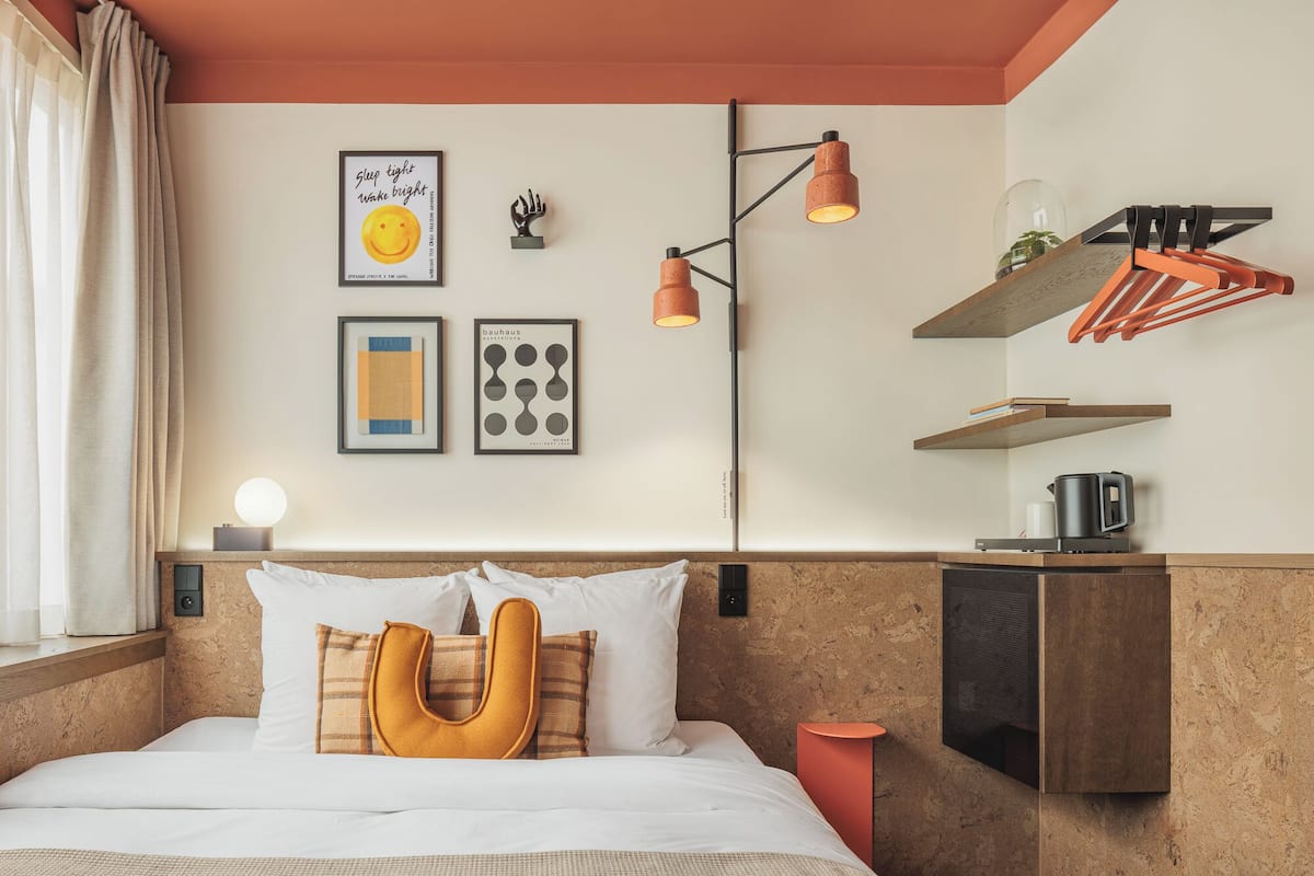 A modern hotel room with a bed, a bedside table with a kettle, a wall-mounted shelf with books, and a unique U-shaped pillow.