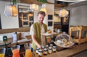 A person serving food at a counter with a variety of pastries and desserts, a glass display case, and a seating area with a sofa and table.