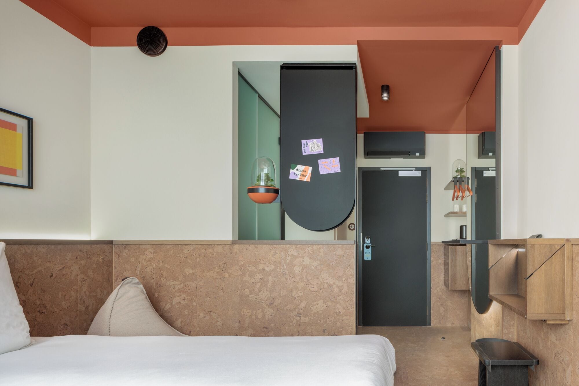 A hotel room with a bed, a small plant, a door, and a mirror.
