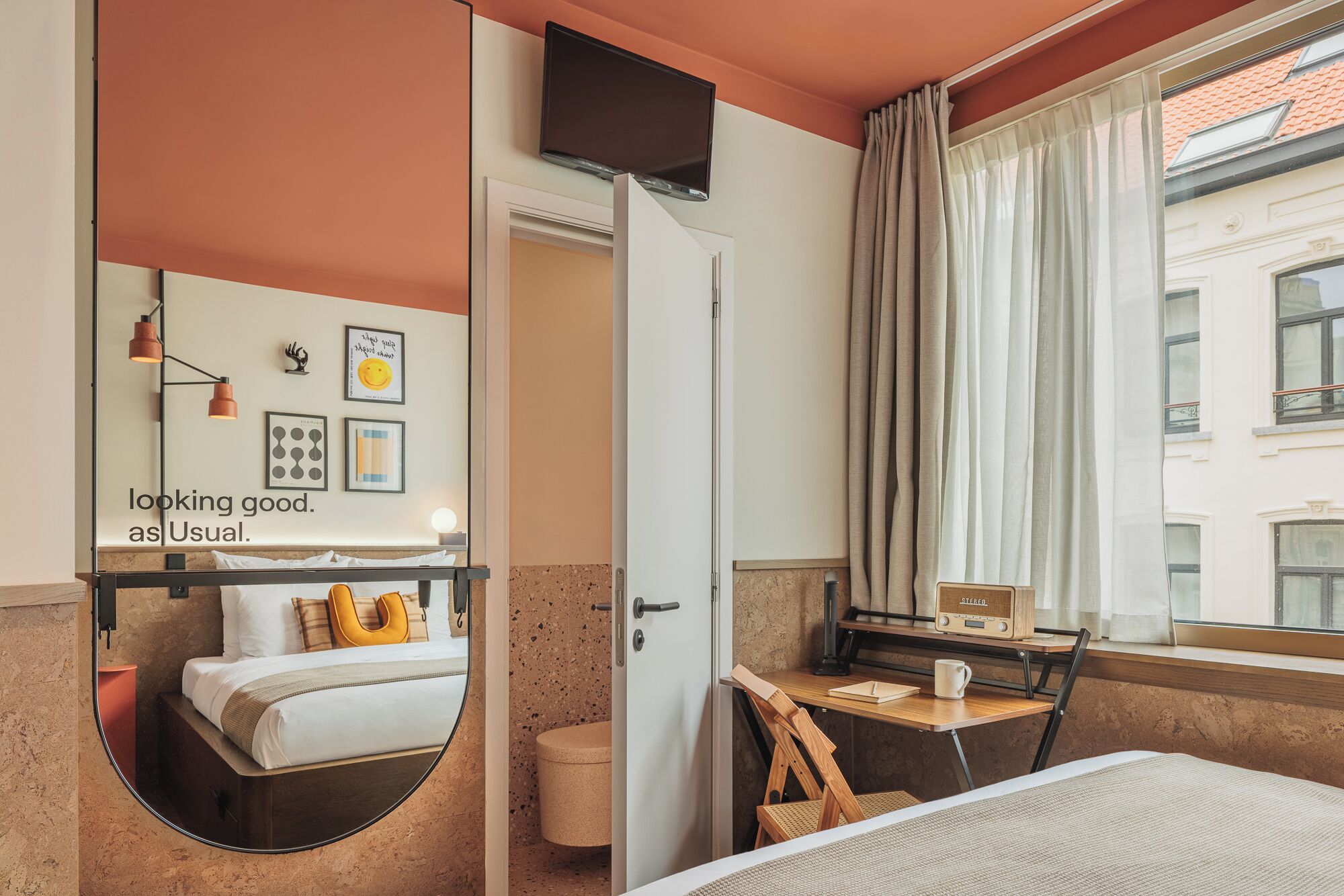Une chambre d’hôtel avec un lit, un bureau avec une radio, un miroir et une fenêtre avec des rideaux.