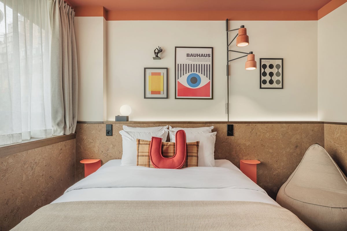 A hotel room with a bed, pillows, a red pillow, a wall-mounted lamp, and framed posters on the wall.