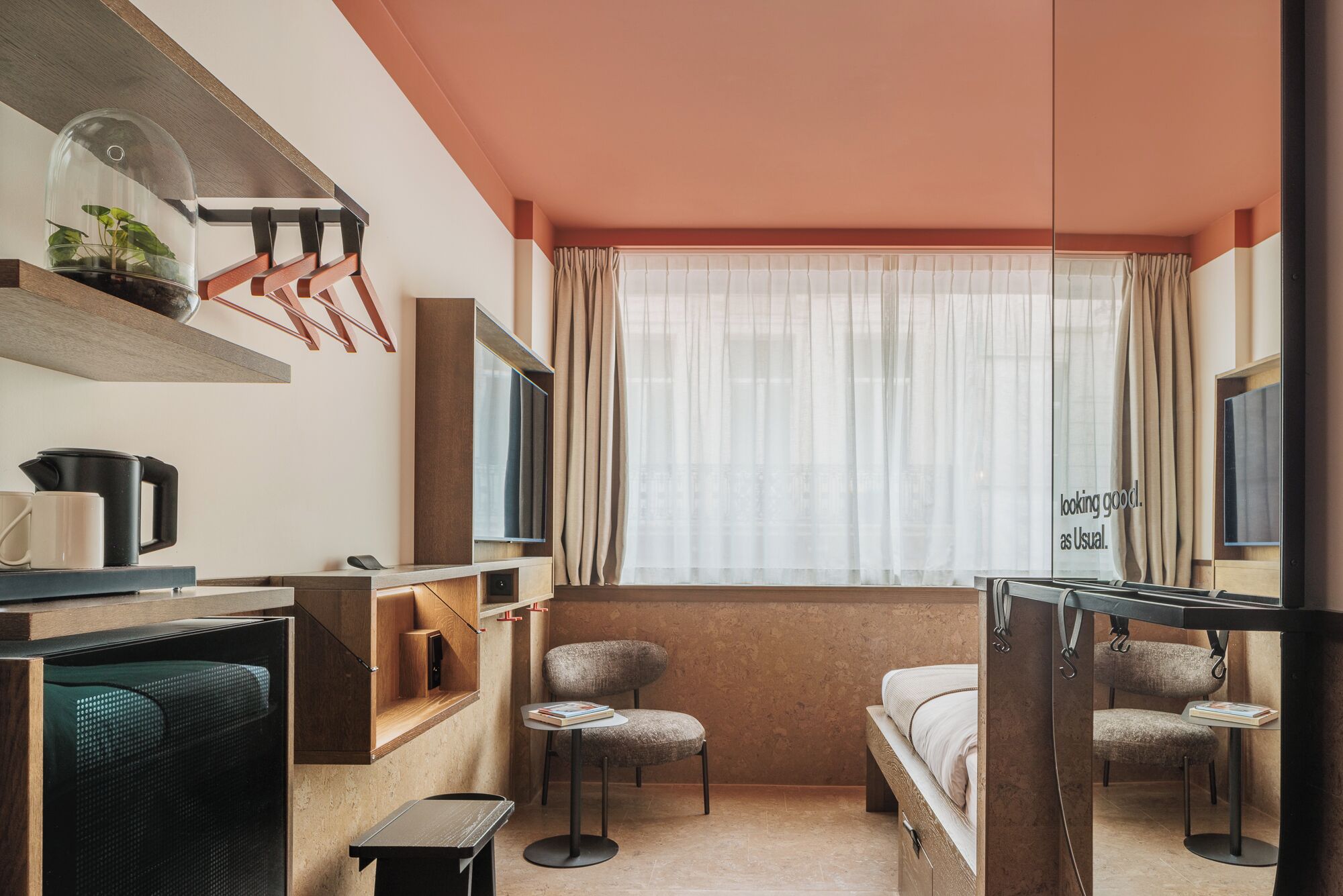 A modern hotel room with a large window, a flat-screen TV, a desk with a chair, a bed, and a small table with a chair.