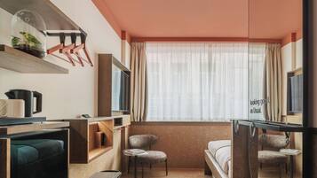 A modern hotel room with a large window, a flat-screen TV, a desk with a chair, a bed, and a small table with a chair.