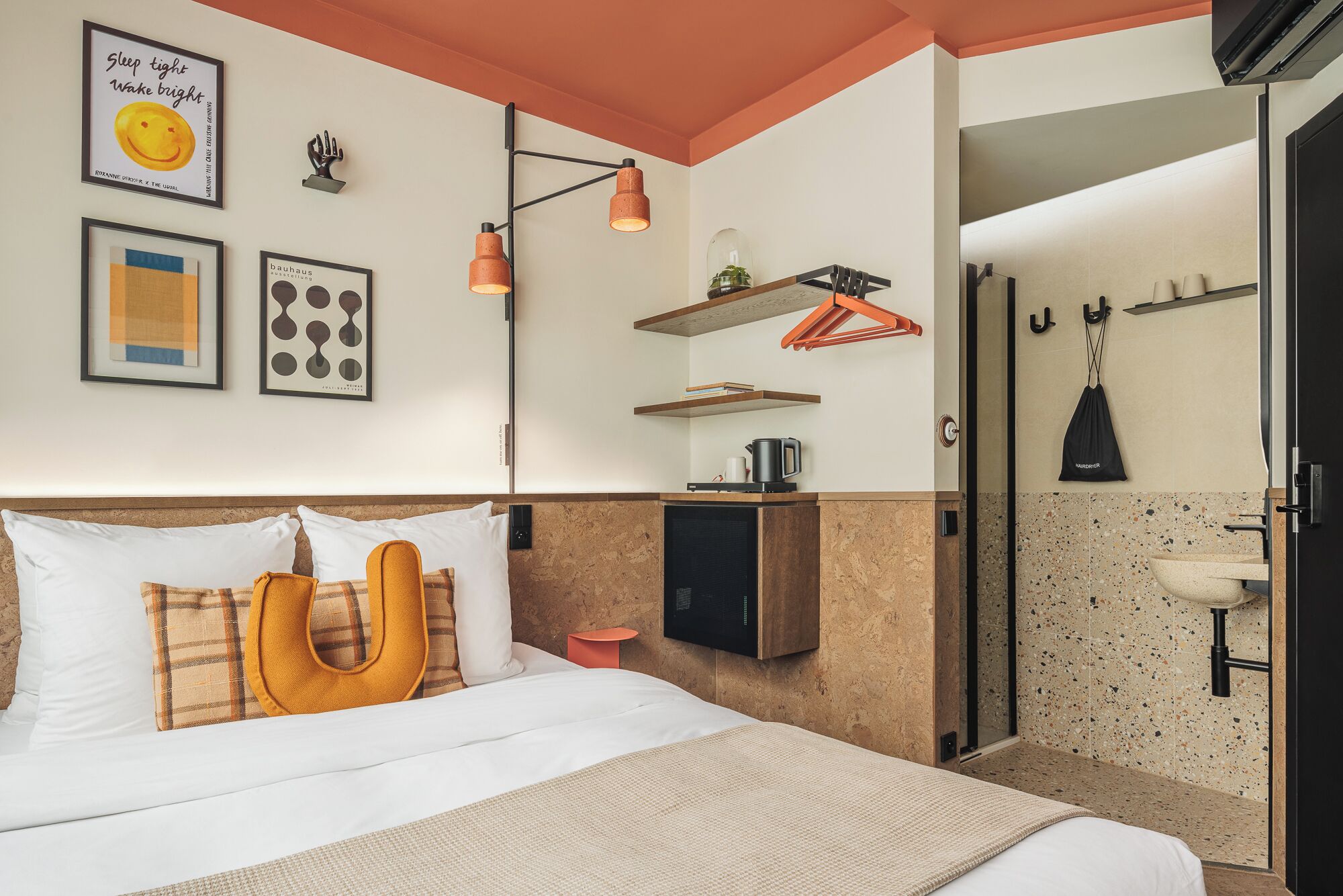 Una camera d'hotel moderna con un letto, una macchina per caffè, un piccolo ripiano con libri e uno spazio doccia.