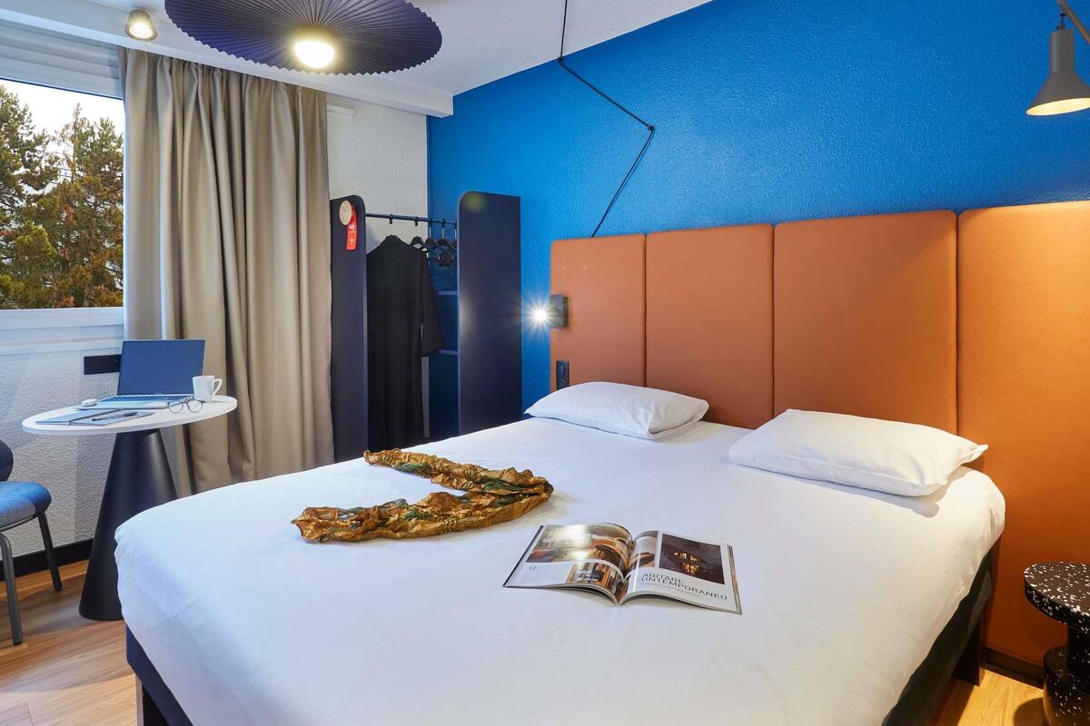 A modern hotel room with a large bed, a small table with a laptop, and a view of trees through the window.