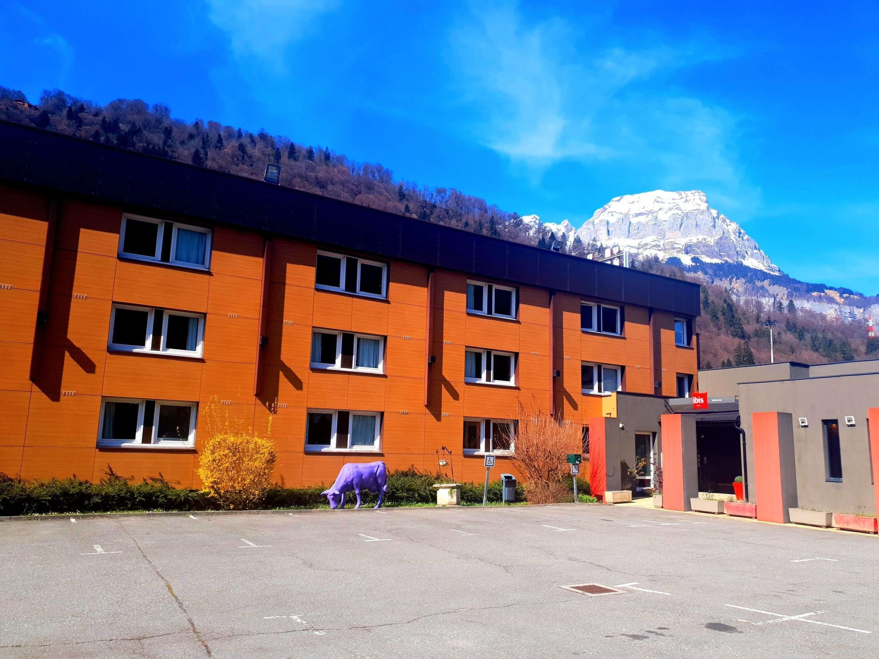 Un edificio de varios pisos con un estacionamiento al frente, una montaña al fondo y una escultura de vaca púrpura.