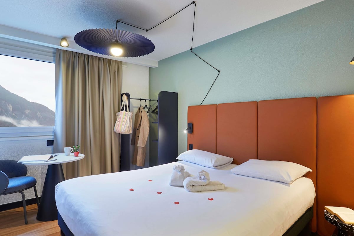 A hotel room with a bed, a desk, a chair, a window with curtains, and a view of the sea.