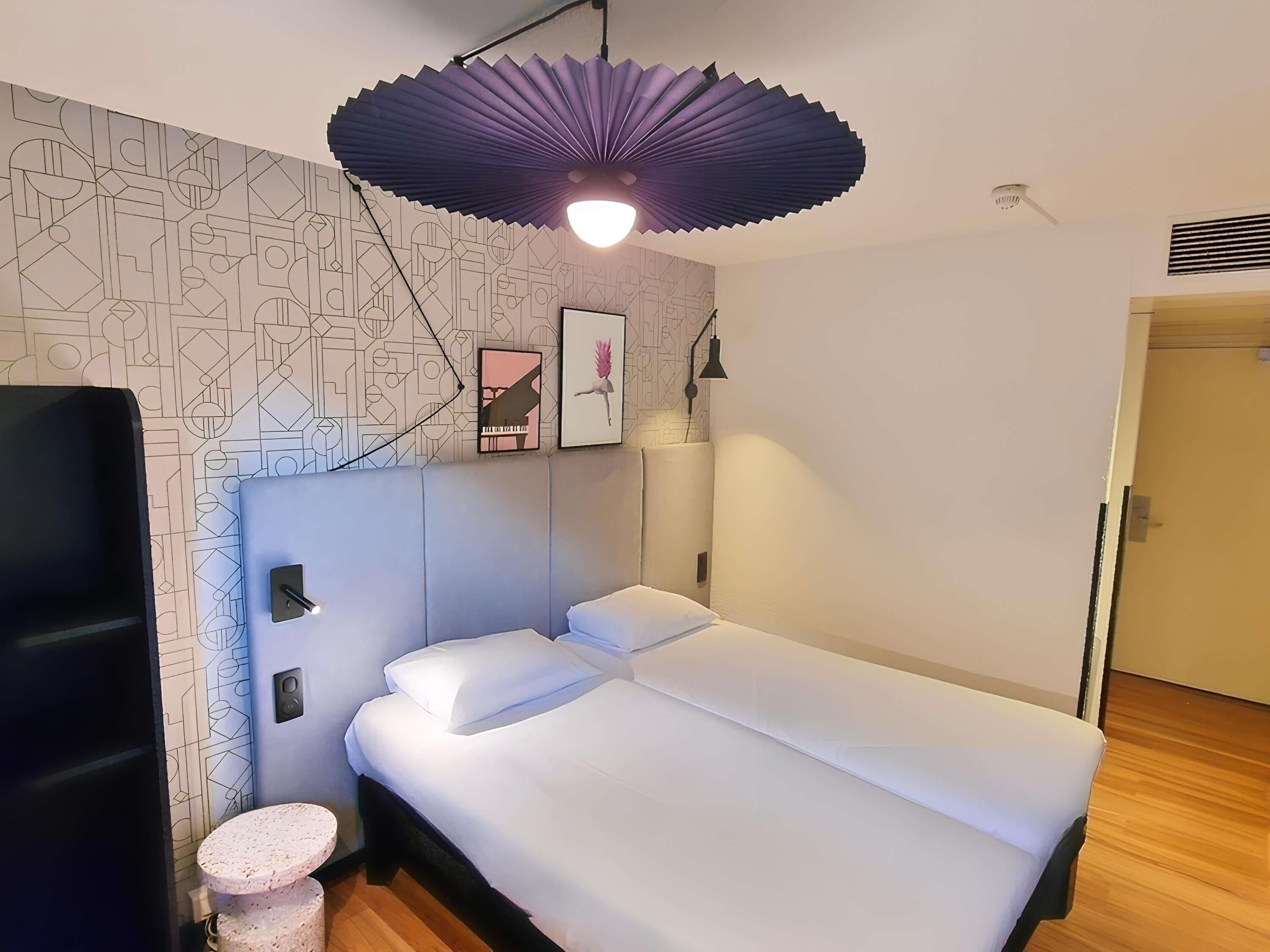 A modern room with a bed, a small round table, a wall-mounted lamp, and geometric-patterned wallpaper.